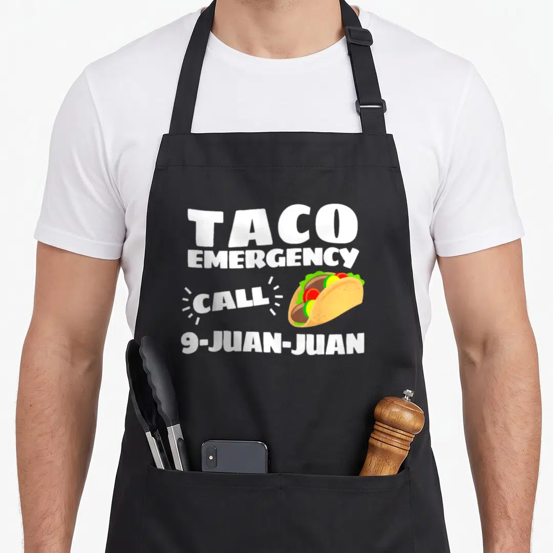 Cinco taco apron - TACO EMERGENCY CALL 9-JUAN-JUAN text next to cartoon taco with lettuce and tomato — shown on a black cotton canvas apron against a white background