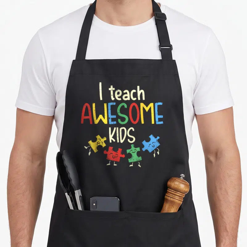 Teach teacher art apron - I teach AWESOME KIDS text with four colorful smiling puzzle pieces in cartoon style — shown on a black cotton canvas apron against a white background