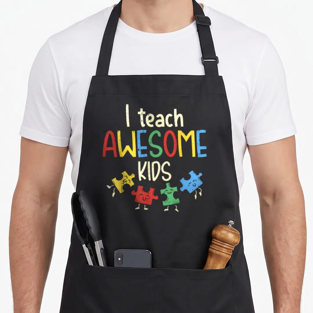 Teach teacher art apron - I teach AWESOME KIDS text with four colorful smiling puzzle pieces in cartoon style — shown on a black cotton canvas apron against a white background