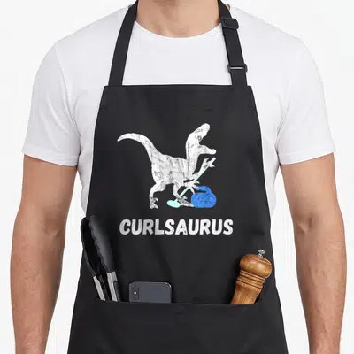 Curling dinosaur apron - Distressed white dinosaur holding blue broom next to blue stone with CURLSAURUS text below — shown on a black cotton canvas apron against a white background