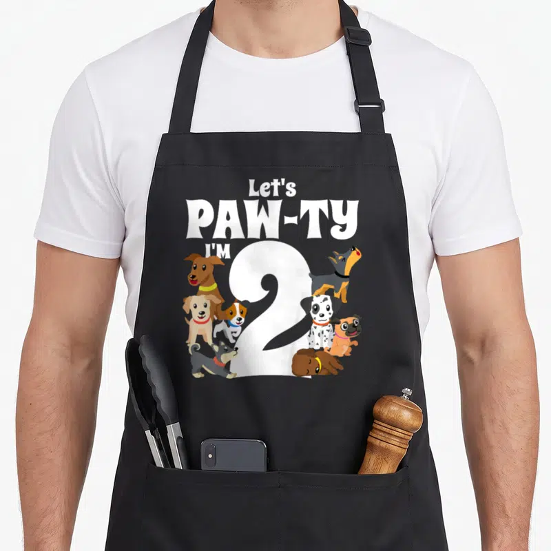 Pawty dog apron - Cartoon dogs surrounding white number 2 with Let's PAW-TY I'M 2 text — shown on a black cotton canvas apron against a white background