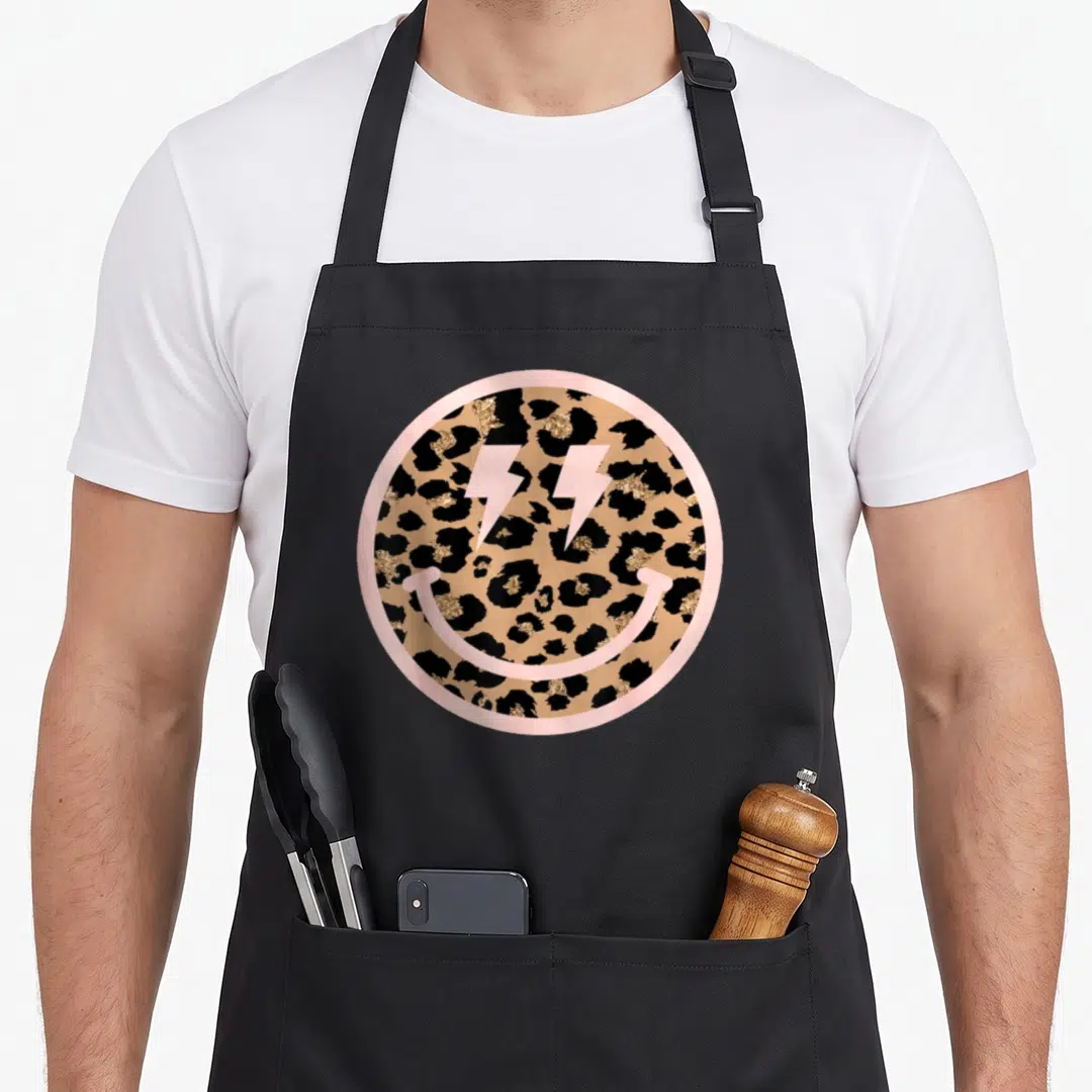 Smile leopard apron - Pink circle with leopard print, pink lightning bolt eyes, and pink smile curve — shown on a black cotton canvas apron against a white background