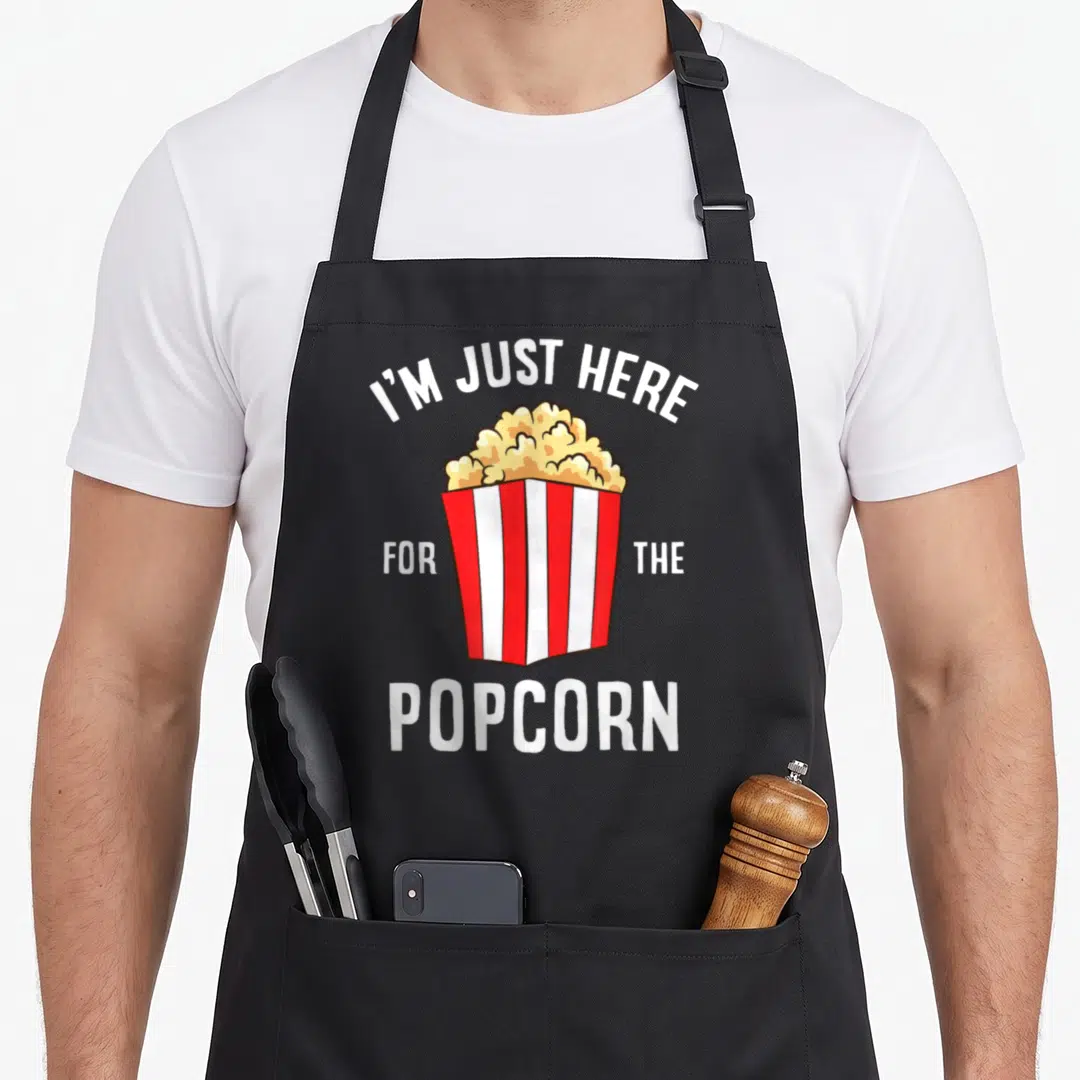 Watching popcorn apron - Striped popcorn bucket with I'M JUST HERE FOR THE POPCORN text — shown on a black cotton canvas apron against a white background