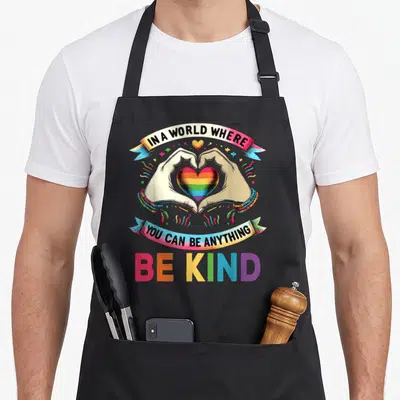 Anything gay pride apron - Hands forming heart around rainbow core, banners with kindness quote, large BE KIND text — shown on a black cotton canvas apron against a white background