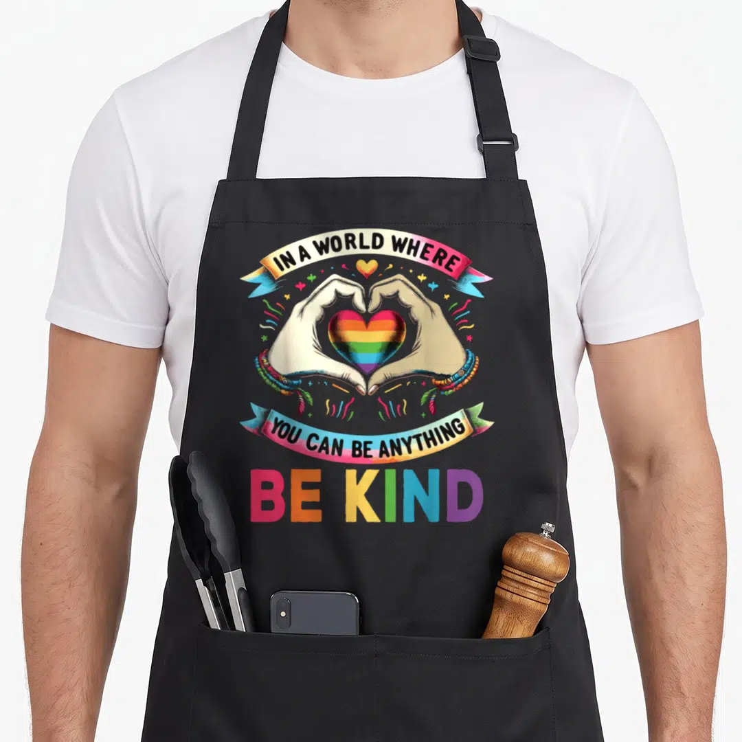 Anything gay pride apron - Hands forming heart around rainbow core, banners with kindness quote, large BE KIND text — shown on a black cotton canvas apron against a white background