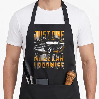 Promise car apron - Distressed orange text Just One More Car I Promise around white muscle car with splatters — shown on a black cotton canvas apron against a white background