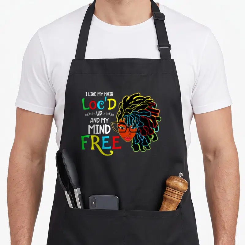 Locd hair apron - Cartoon woman with colorful locs next to stacked text I Like My Hair Loc'd Up and My Mind Free — shown on a black cotton canvas apron against a white background