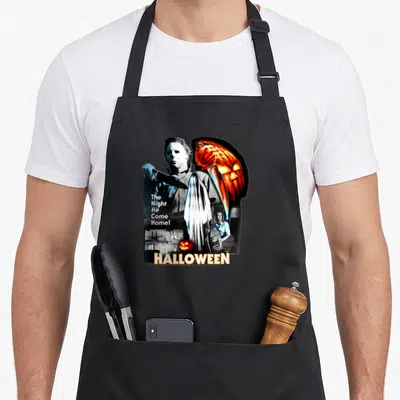 Night horror movie apron - Michael Myers collage with knife pumpkin and The Night He Came Home text — shown on a black cotton canvas apron against a white background