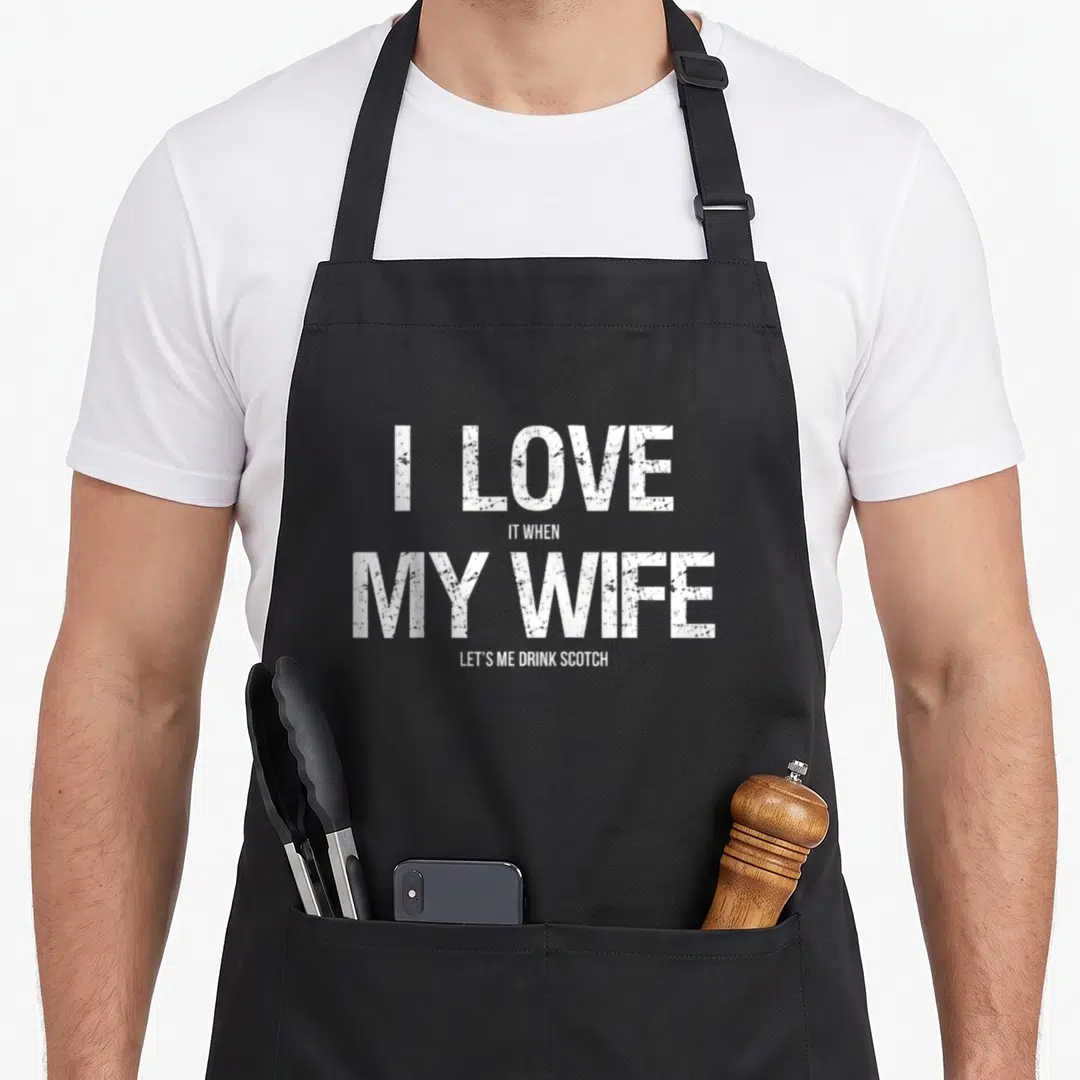 Scotch wife apron - White distressed text reading I LOVE IT WHEN MY WIFE LET'S ME DRINK SCOTCH stacked in four lines — shown on a black cotton canvas apron against a white background