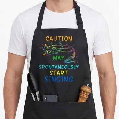 Caution singing bird apron - Stacked colorful text reading caution may spontaneously start singing with rainbow musical — shown on a black cotton canvas apron against a white background