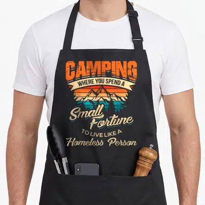 Camp happy camper apron - Camping Where You Spend a Small Fortune To Live Like A Homeless Person text over retro — shown on a black cotton canvas apron against a white background