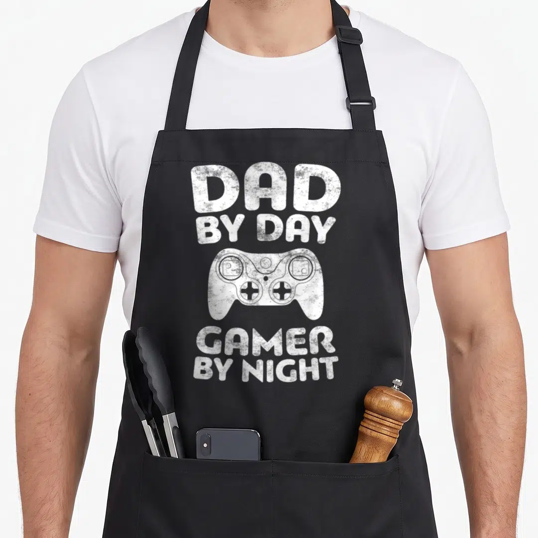 Gamer father day apron - DAD BY DAY GAMER BY NIGHT text with a game controller illustration in white distressed print — shown on a black cotton canvas apron against a white background