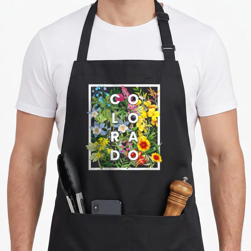 Coloradan pride apron - White COLORADO text over a dense cluster of blue, yellow, red, and green wildflowers in a white — shown on a black cotton canvas apron against a white background