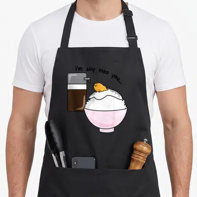 Soy egg apron - Cartoon rice bowl with sauce bottle and i'm soy into you text — shown on a black cotton canvas apron against a white background