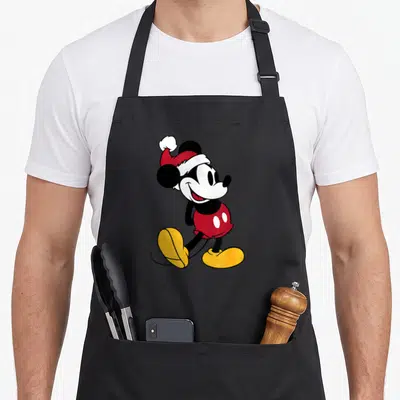 Funny holiday apron - Cartoon Mickey Mouse in red Santa hat and shorts with yellow shoes — shown on a black cotton canvas apron against a white background