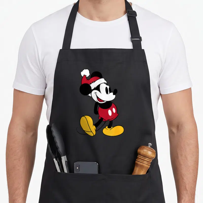 Funny holiday apron - Cartoon Mickey Mouse in red Santa hat and shorts with yellow shoes — shown on a black cotton canvas apron against a white background