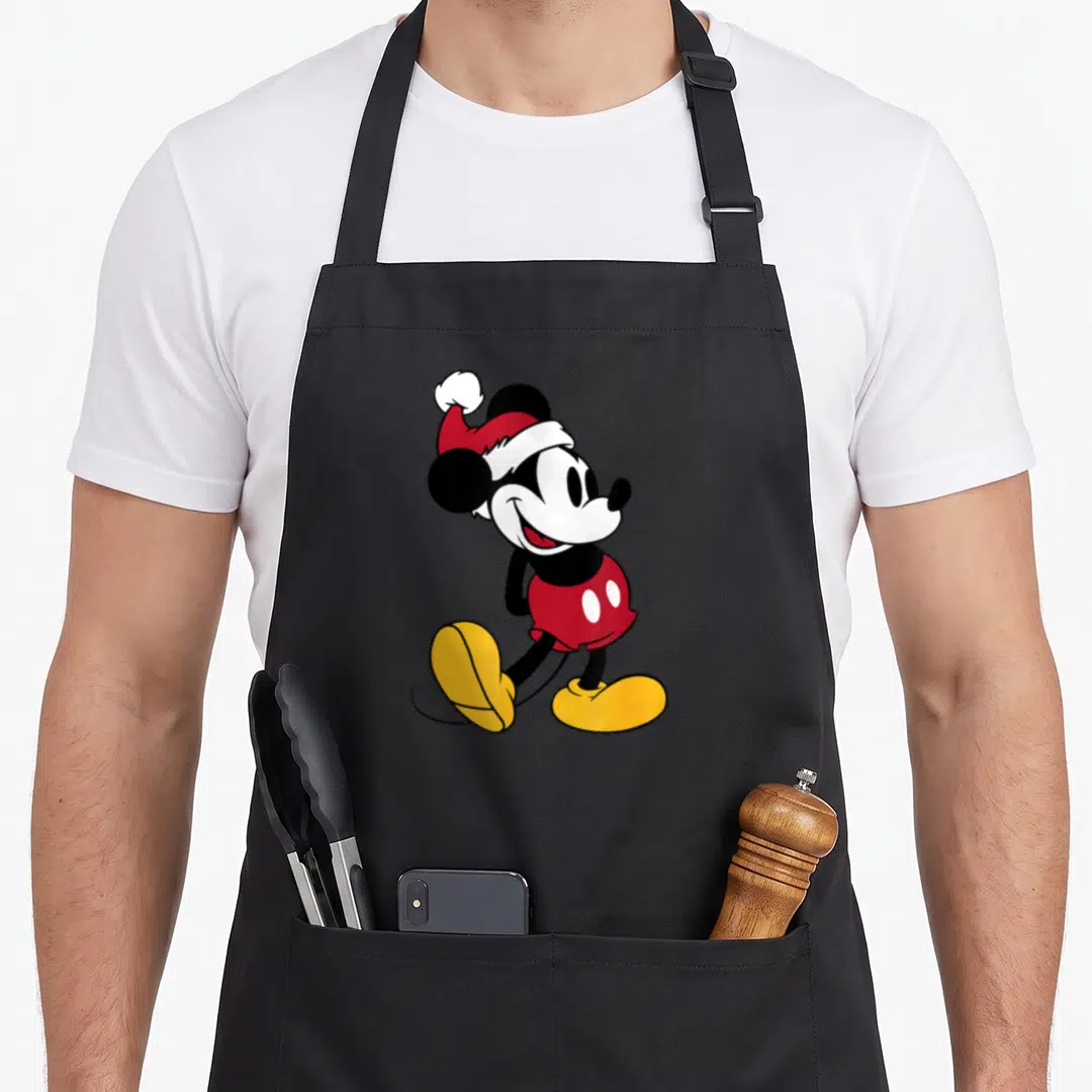 Funny holiday apron - Cartoon Mickey Mouse in red Santa hat and shorts with yellow shoes — shown on a black cotton canvas apron against a white background