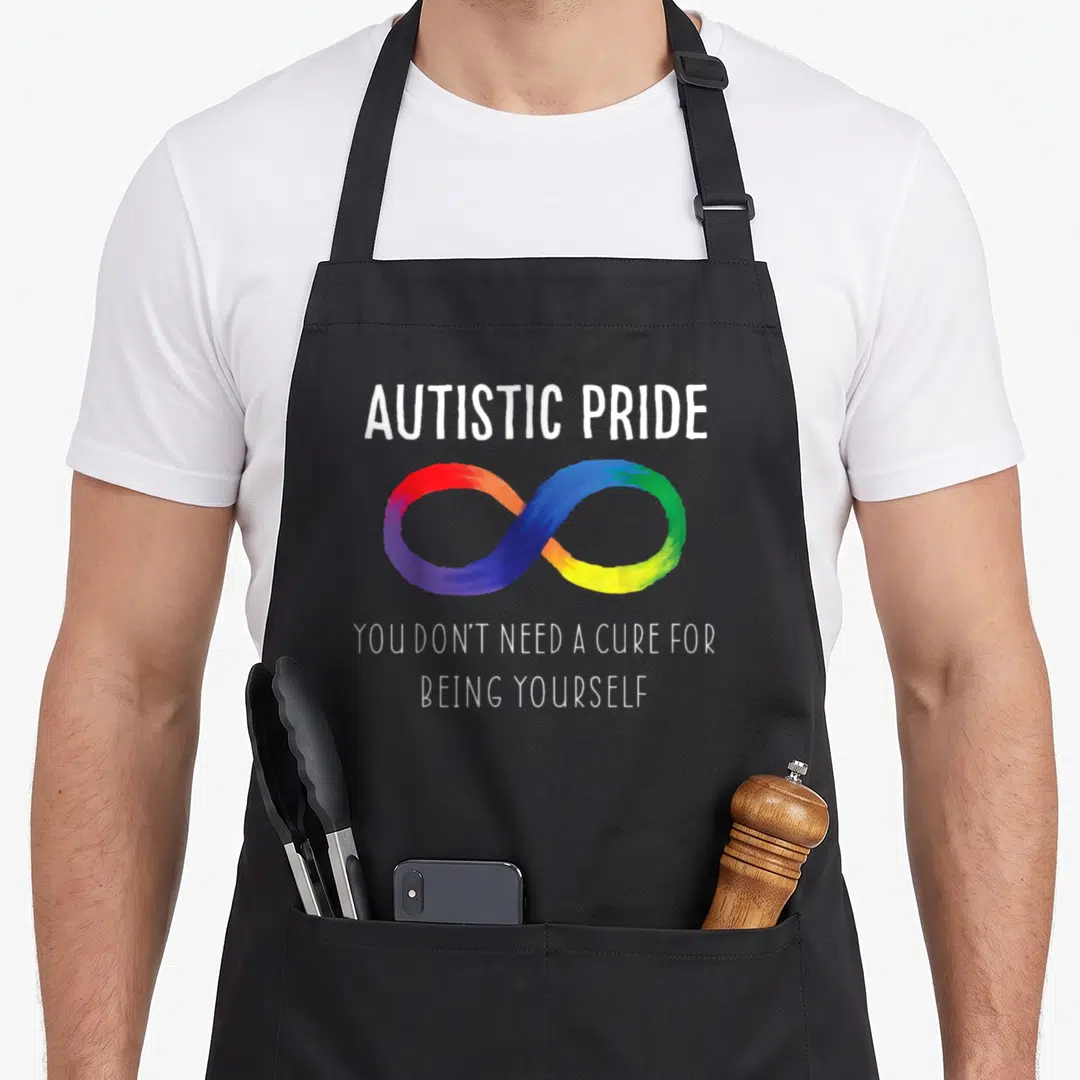 Autistic pride apron - Rainbow infinity symbol with white text reading autistic pride and you dont need a cure — shown on a black cotton canvas apron against a white background