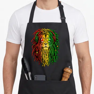 Sunglass lion apron - Cartoon lion head with red yellow green dreadlocks sunglasses and headphones — shown on a black cotton canvas apron against a white background