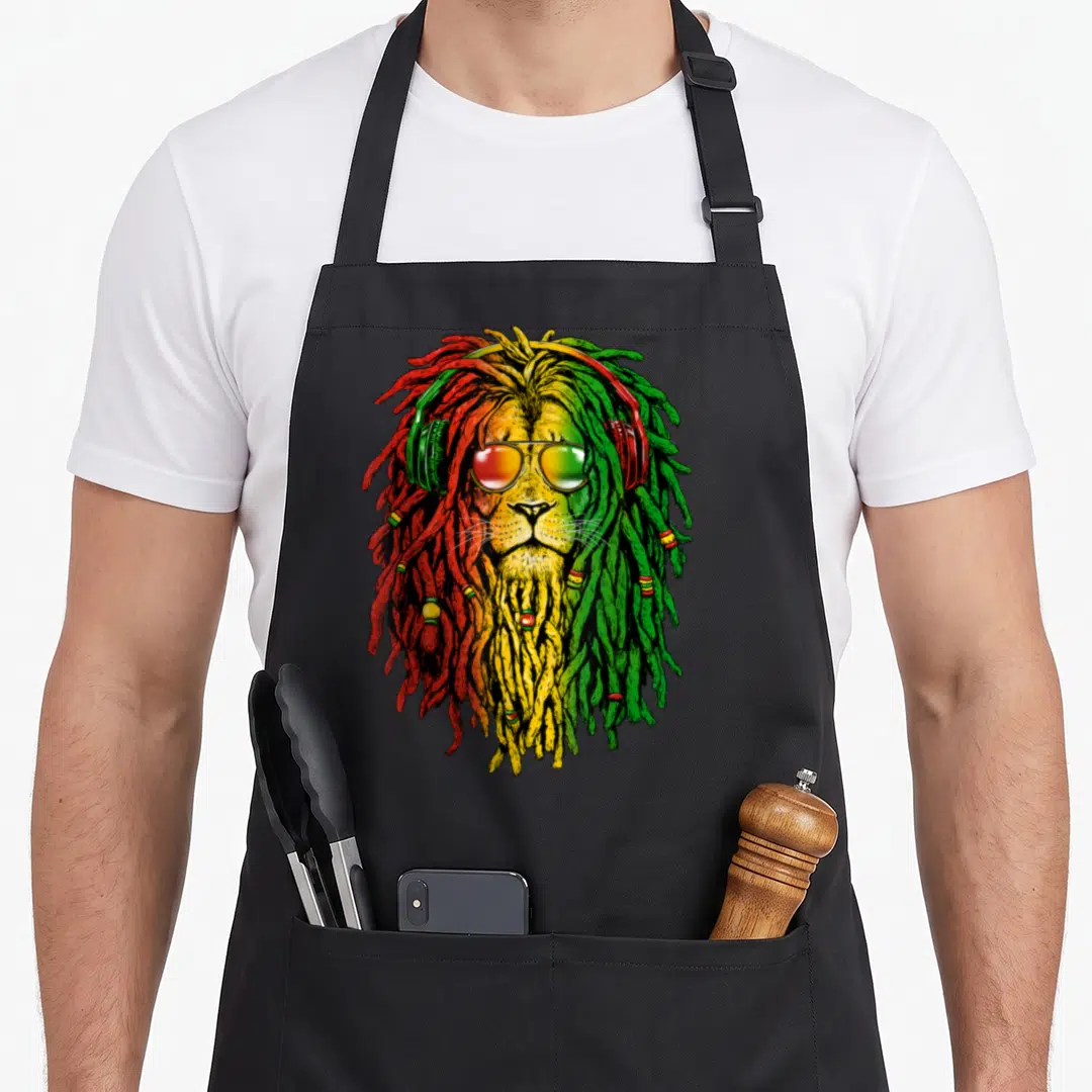 Sunglass lion apron - Cartoon lion head with red yellow green dreadlocks sunglasses and headphones — shown on a black cotton canvas apron against a white background