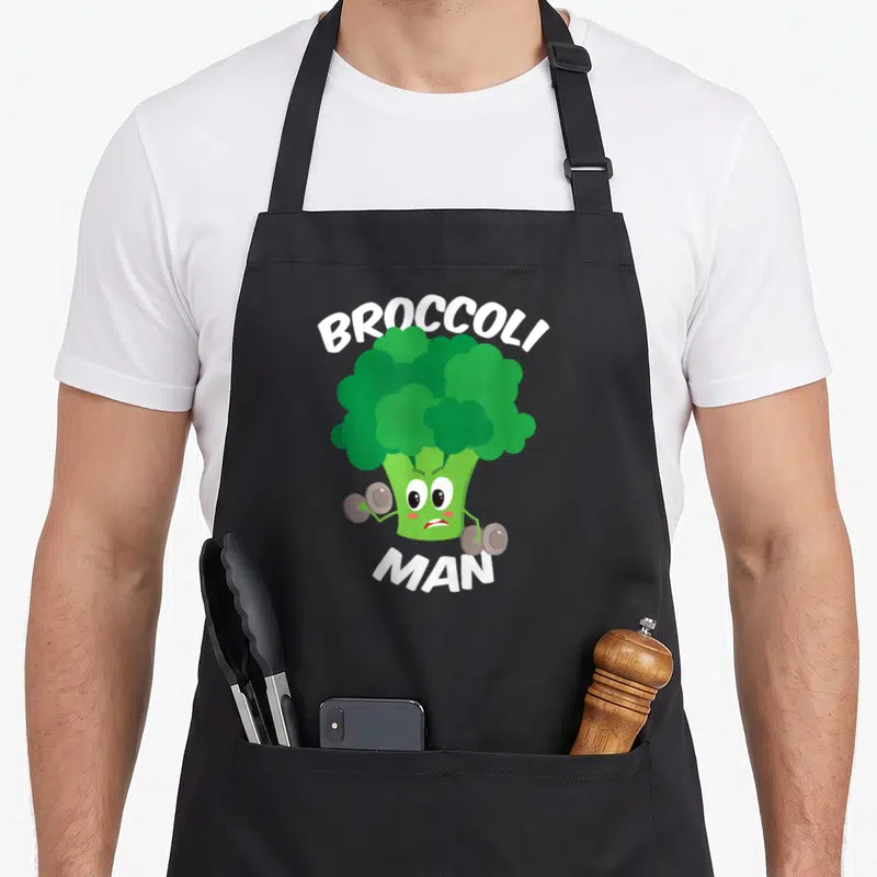 Funny broccoli vegetable apron - Cartoon green broccoli lifting grey dumbbells with white BROCCOLI MAN text — shown on a black cotton canvas apron against a white background