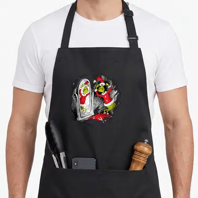 Saint holiday apron - Grinch in red suit looking at mirror reflection with scissors and fabric on floor — shown on a black cotton canvas apron against a white background