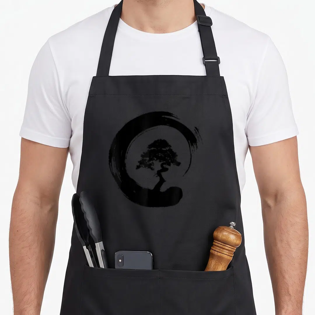 Enso tree apron - Black line art bonsai tree inside a brush stroke enso circle — shown on a black cotton canvas apron against a white background