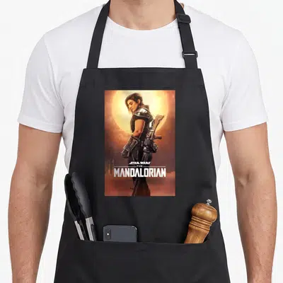 Poster dunes apron - Cara Dune in armor against yellow sun with white Star Wars The Mandalorian text — shown on a black cotton canvas apron against a white background