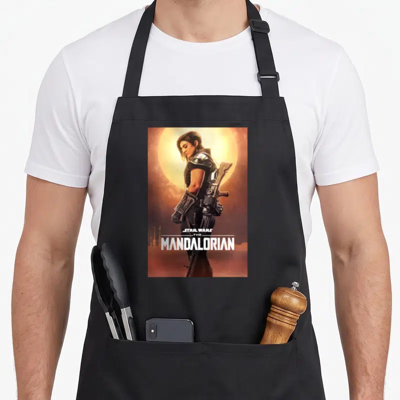 Poster dunes apron - Cara Dune in armor against yellow sun with white Star Wars The Mandalorian text — shown on a black cotton canvas apron against a white background