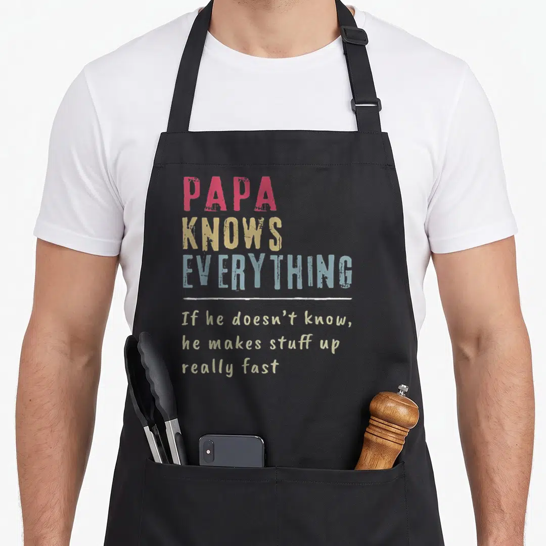 Cute papa apron - PAPA KNOWS EVERYTHING text in red yellow blue with white line separator — shown on a black cotton canvas apron against a white background
