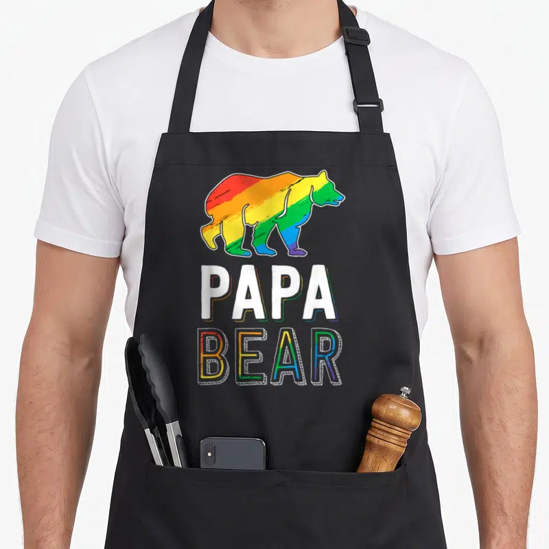 Lgbtq bear apron - Rainbow cartoon bear above white PAPA and outlined BEAR text — shown on a black cotton canvas apron against a white background