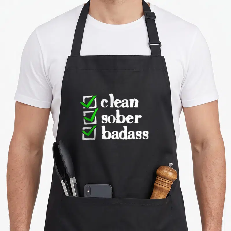 Badass clean apron - White checklist with three green checkmarks beside text reading clean sober badass in distressed — shown on a black cotton canvas apron against a white background
