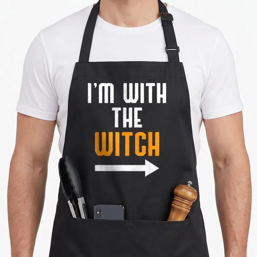 Funny halloween witch apron - Stacked white and orange text reading I'M WITH THE WITCH with a white arrow pointing right — shown on a black cotton canvas apron against a white background