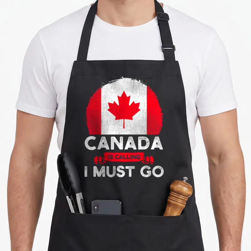 Calling canada aprons - Distressed circular Canadian flag with red ribbon text reading Canada Is Calling I Must Go — shown on a black cotton canvas apron against a white background