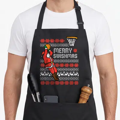 Swishmas xmas apron - Santa jumping with basketball next to Merry Swishmas text on knit pattern — shown on a black cotton canvas apron against a white background