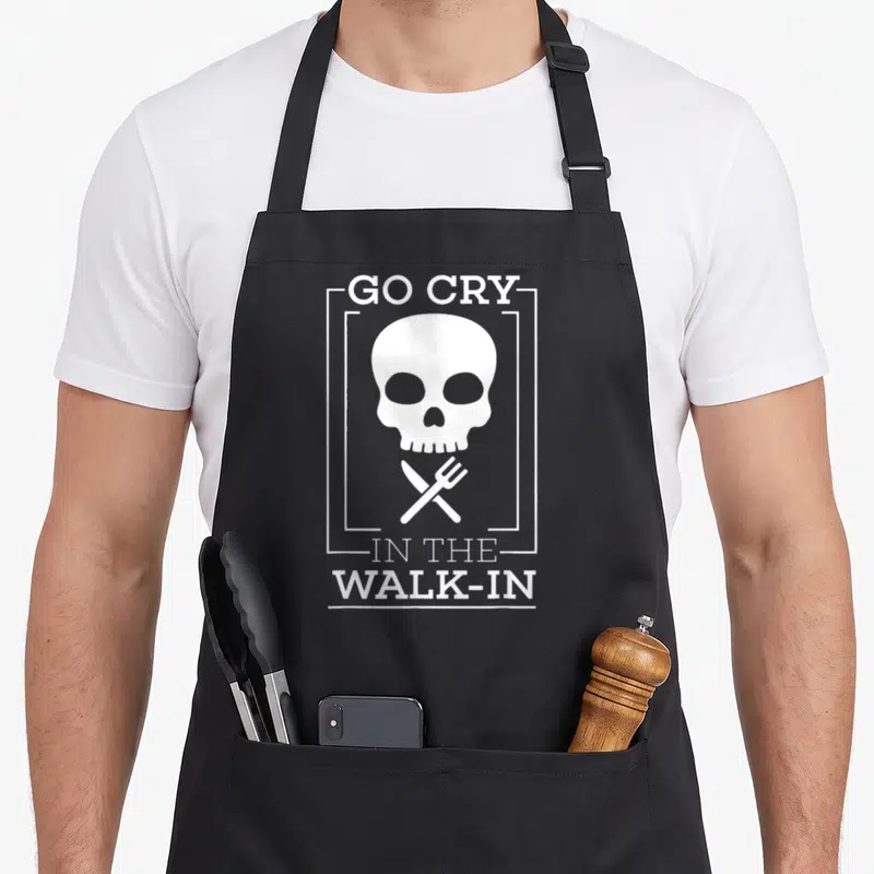 Funny line apron - White skull inside rectangular frame with crossed knife and fork and GO CRY IN THE WALK-IN text — shown on a black cotton canvas apron against a white background