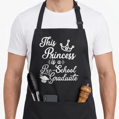 Pre school graduation apron - White cursive text reading This Princess is a Pre-School Graduate with crown and cap icons — shown on a black cotton canvas apron against a white background