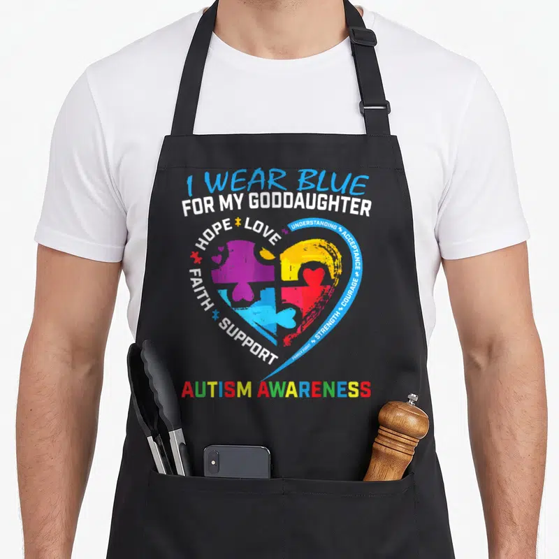 Blue autism acceptance apron - Puzzle heart in blue white yellow red purple with I WEAR BLUE FOR MY GODDAUGHTER text — shown on a black cotton canvas apron against a white background
