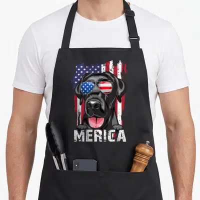 Retriever 4th of july apron - Black dog with flag sunglasses and MERICA text on distressed flag background — shown on a black cotton canvas apron against a white background