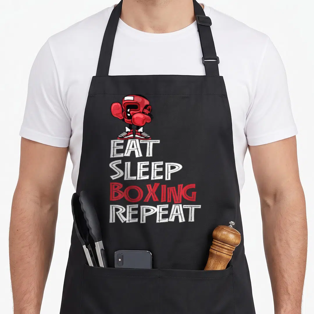 Sleep boxing apron - Cartoon boxer standing on stacked EAT SLEEP BOXING REPEAT text in red and white — shown on a black cotton canvas apron against a white background