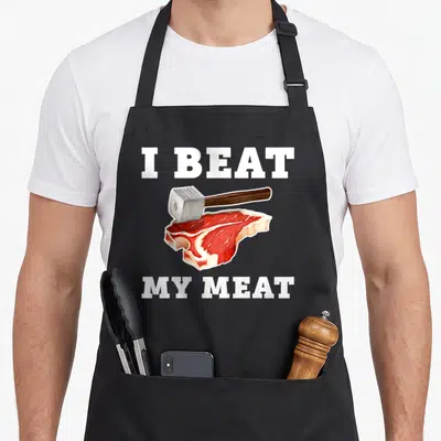 Hammer meat aprons - I Beat My Meat text with silver mallet hitting raw red steak in realistic style — shown on a black cotton canvas apron against a white background