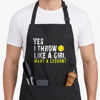Cute softball apron - Stacked white text Yes I Throw Like A Girl Want A Lesson next to yellow softball with red — shown on a black cotton canvas apron against a white background