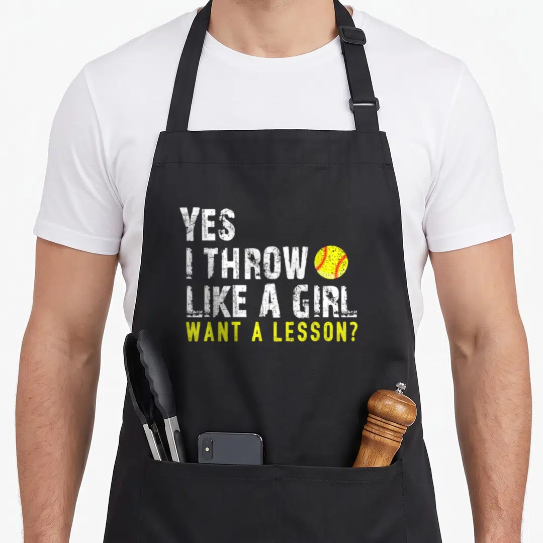 Cute softball apron - Stacked white text Yes I Throw Like A Girl Want A Lesson next to yellow softball with red — shown on a black cotton canvas apron against a white background
