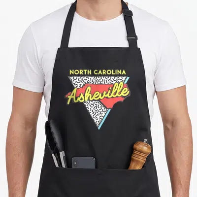 Asheville 80s apron - Retro red NC map with yellow Asheville script over a black and white squiggle triangle — shown on a black cotton canvas apron against a white background