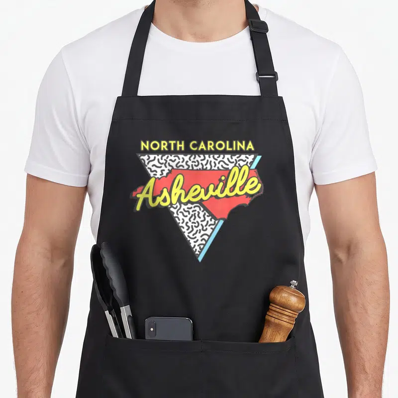 Asheville 80s apron - Retro red NC map with yellow Asheville script over a black and white squiggle triangle — shown on a black cotton canvas apron against a white background