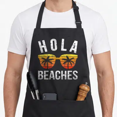 Hola summer aprons - Retro sunglasses with yellow orange red sunset and two palm trees below Hola Beaches text — shown on a black cotton canvas apron against a white background