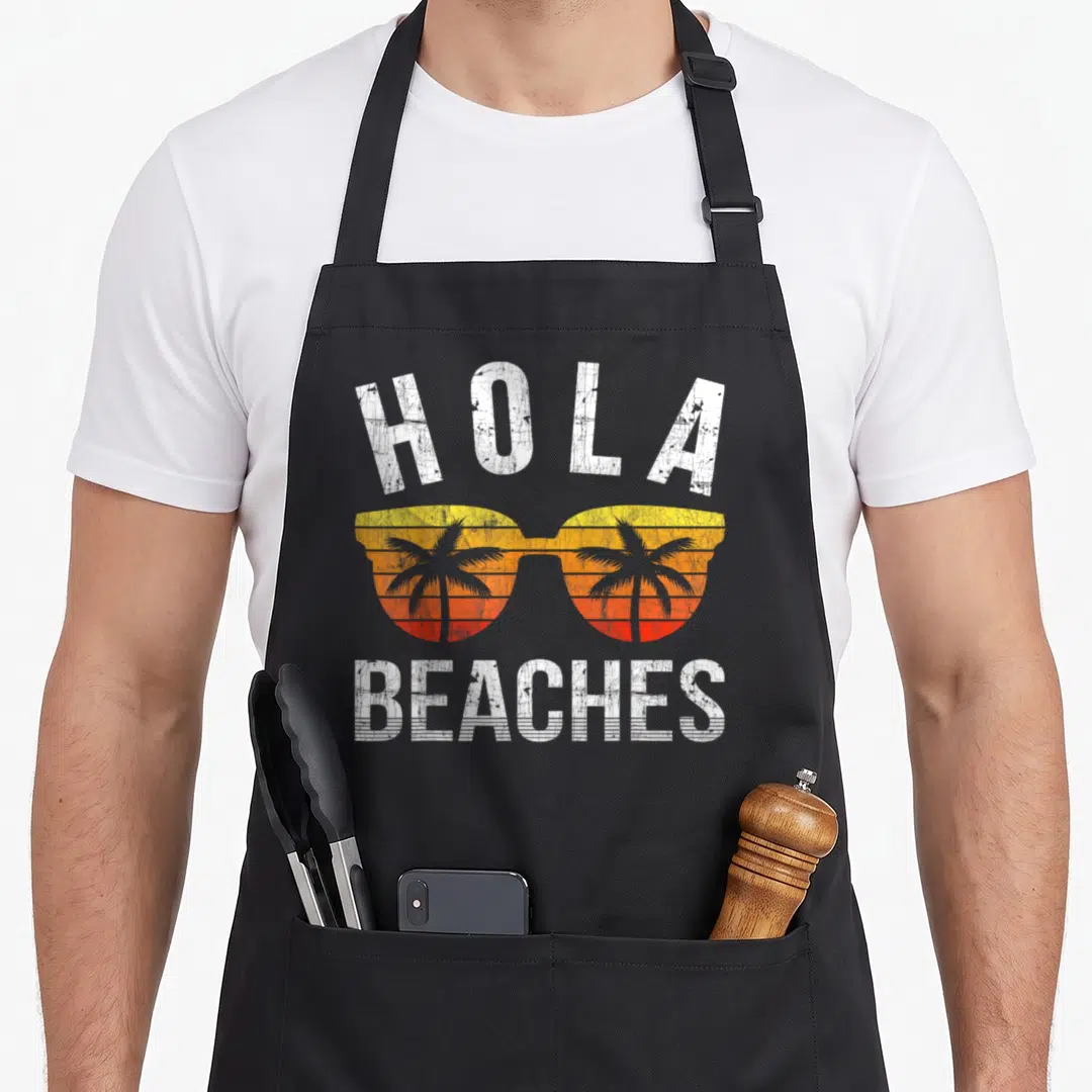 Hola summer aprons - Retro sunglasses with yellow orange red sunset and two palm trees below Hola Beaches text — shown on a black cotton canvas apron against a white background