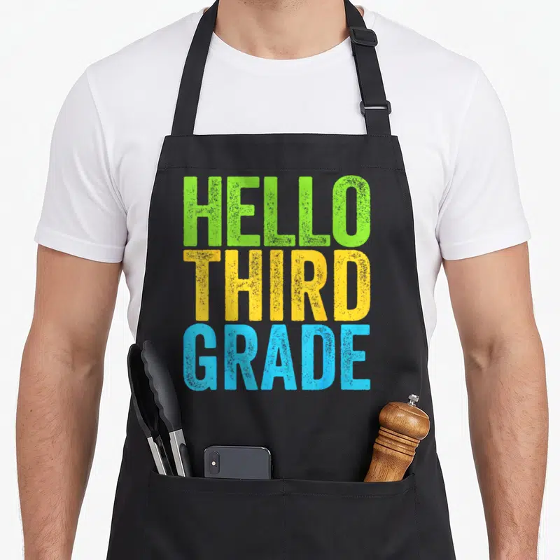 Hello third teacher apron - Stacked HELLO THIRD GRADE text in green, yellow, and blue distressed block letters — shown on a black cotton canvas apron against a white background