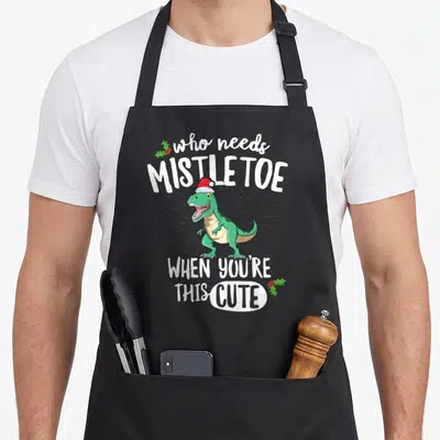 Rex christmas apron - Green T-Rex Santa hat Who Needs Mistletoe When You're This Cute text holly — shown on a black cotton canvas apron against a white background