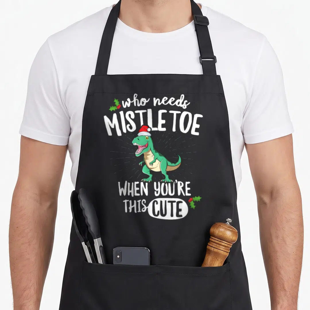 Rex christmas apron - Green T-Rex Santa hat Who Needs Mistletoe When You're This Cute text holly — shown on a black cotton canvas apron against a white background